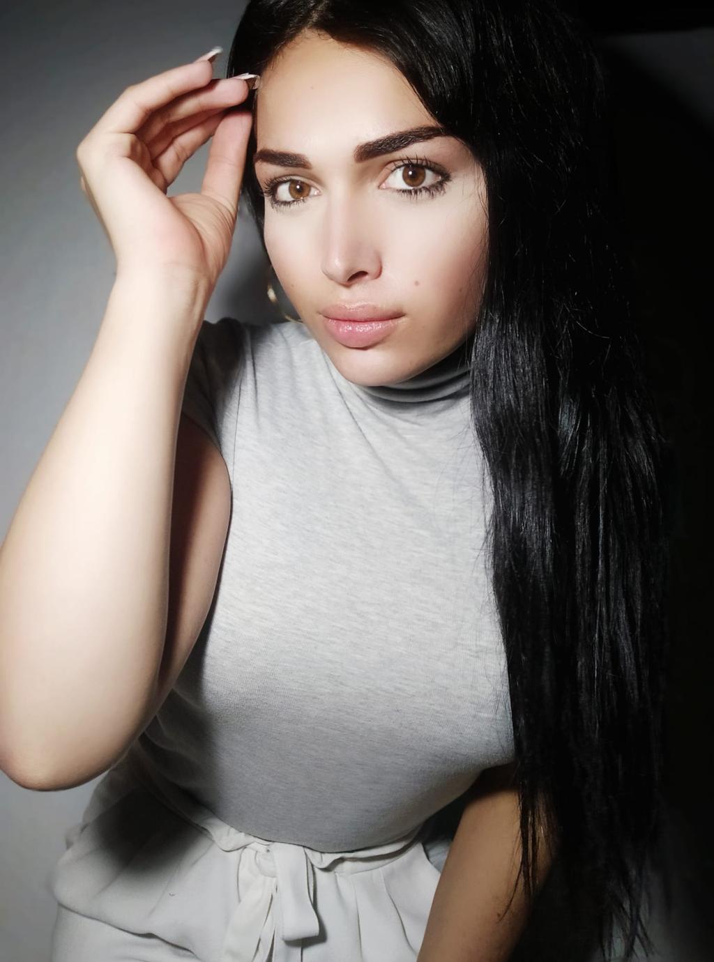 Escort trans en Santander con cabello largo y lacio, luciendo una blusa gris de manga corta y una pose relajada.