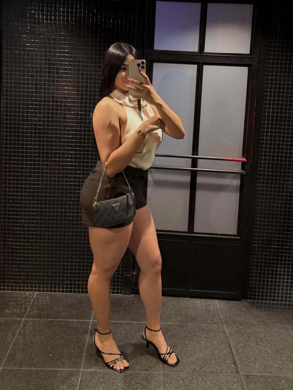 Escort en Pamplona luciendo una blusa blanca y shorts oscuros, posando con una bolsa y sandalias de tacón.