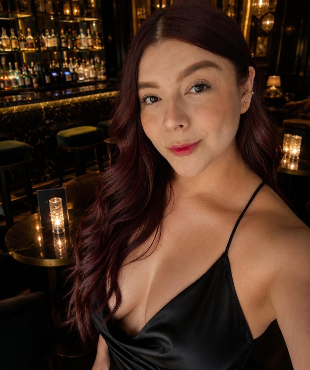 Escort en Gijón con cabello castaño oscuro y vestido negro, posando en un bar elegante con luz tenue.