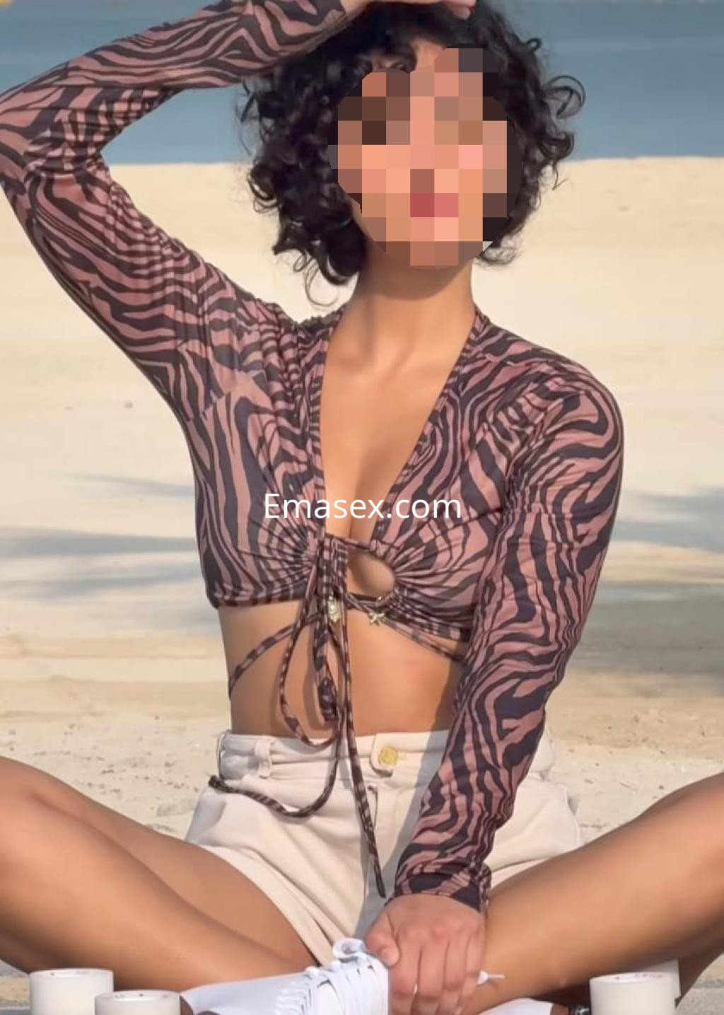 Puta con blusa de estampado animal y pantalones cortos, sentada en la playa de Santiago, mostrando una pose relajada.