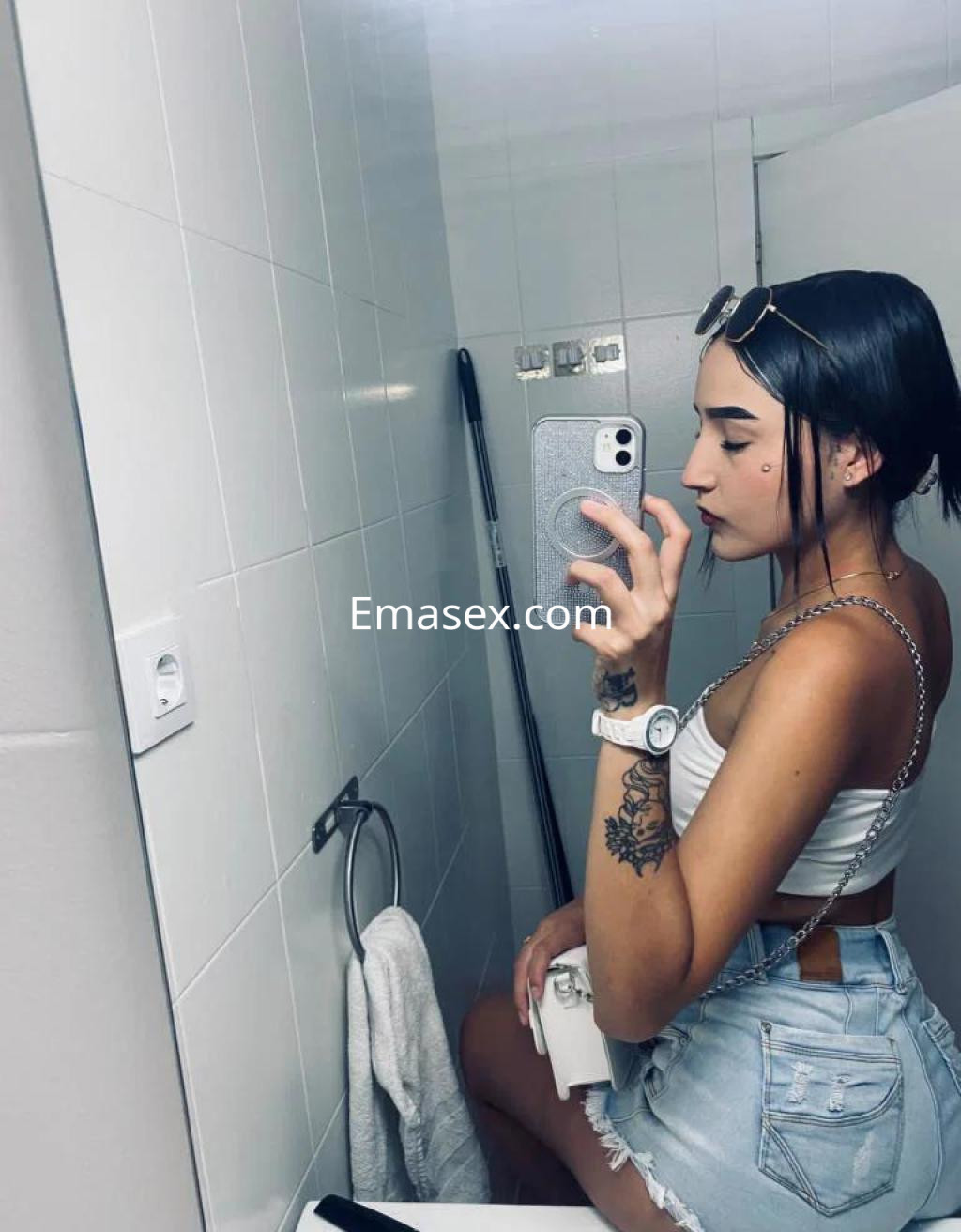 Escort en Cáceres, posando en el baño con top blanco, shorts de mezclilla y gafas de sol.
