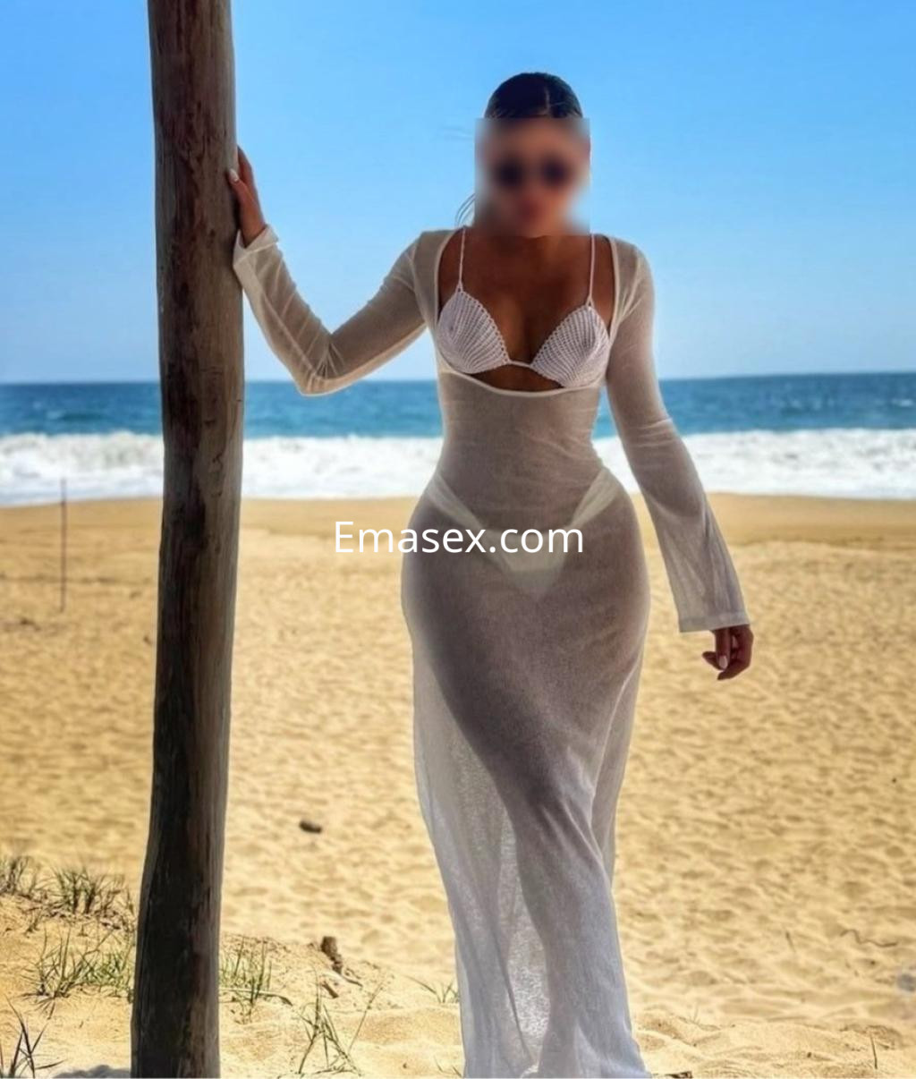 Escort en Santiago con vestido blanco transparente, de pie en la playa, con el mar y el cielo azul de fondo.