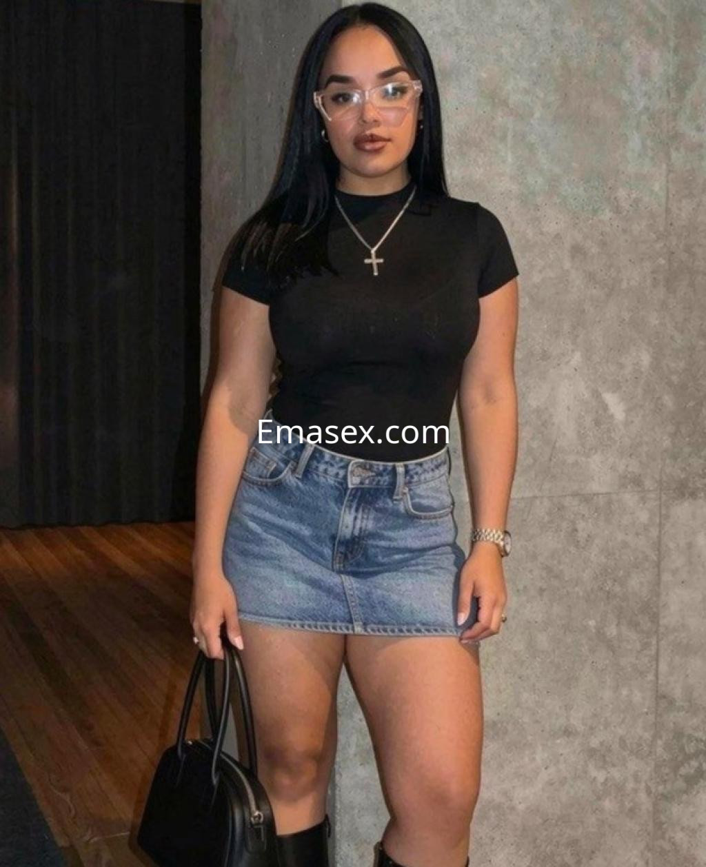 La puta viste una camiseta negra ajustada y una falda de mezclilla, posando en un entorno moderno en Málaga.