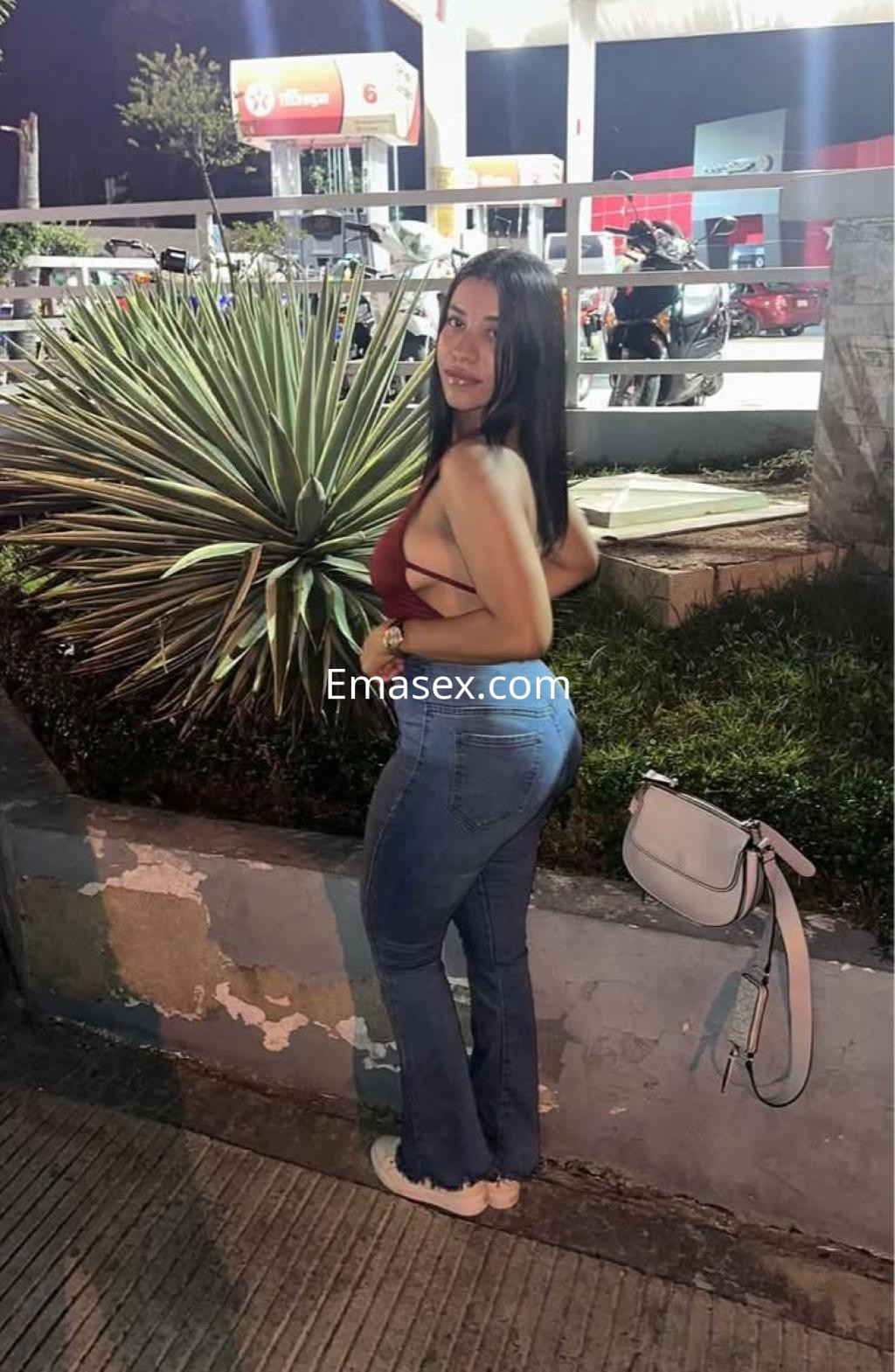 Puta en Málaga con top rojo y jeans acampanados, posando sonriente junto a plantas en un entorno urbano nocturno.