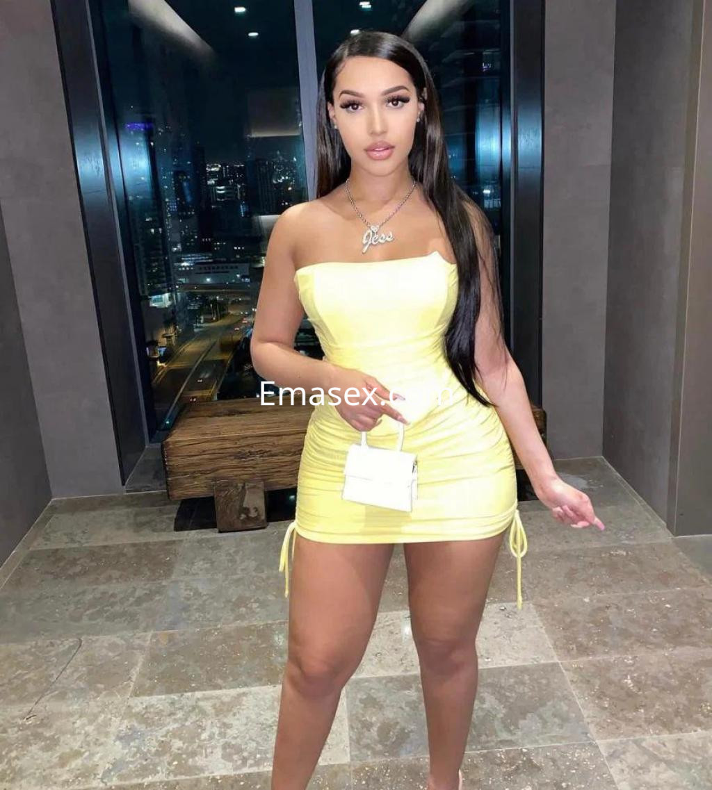 La escort en Albacete viste un vestido amarillo ajustado y sostiene un pequeño bolso blanco, sonriendo en un entorno urbano.