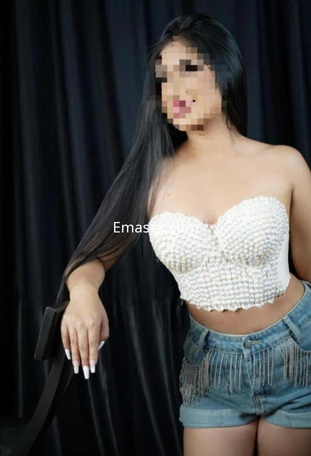 Escort en Badajoz con cabello largo y lacio, viste un top blanco y shorts de mezclilla, posando con confianza.
