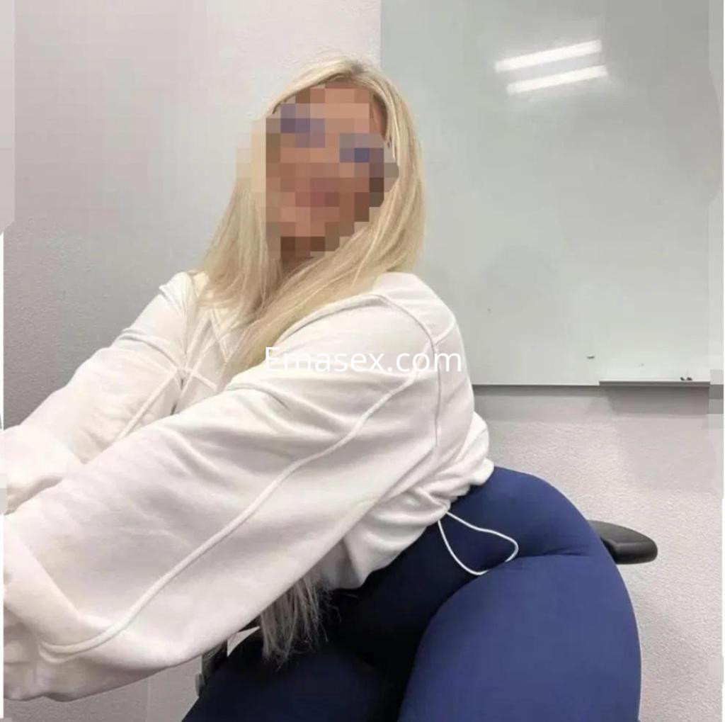 Escort en Cáceres con cabello rubio largo, vistiendo sudadera blanca y leggings azules, sentada en una silla.