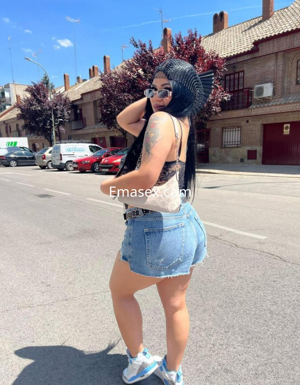 Escort en Madrid luciendo un top negro, shorts de mezclilla y gafas de sol, posando en una calle soleada.