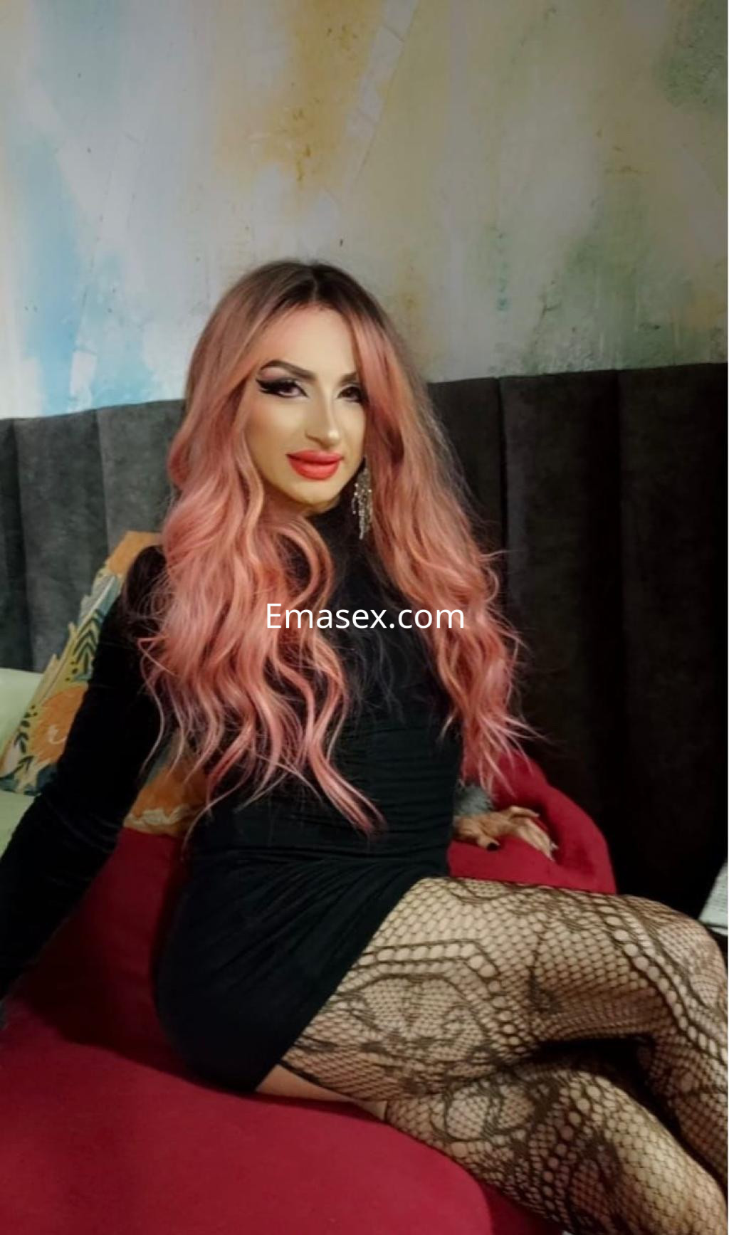 Escort trans en Irún con cabello largo y rosa, luciendo un vestido negro y medias de red, sentada en un sofá.