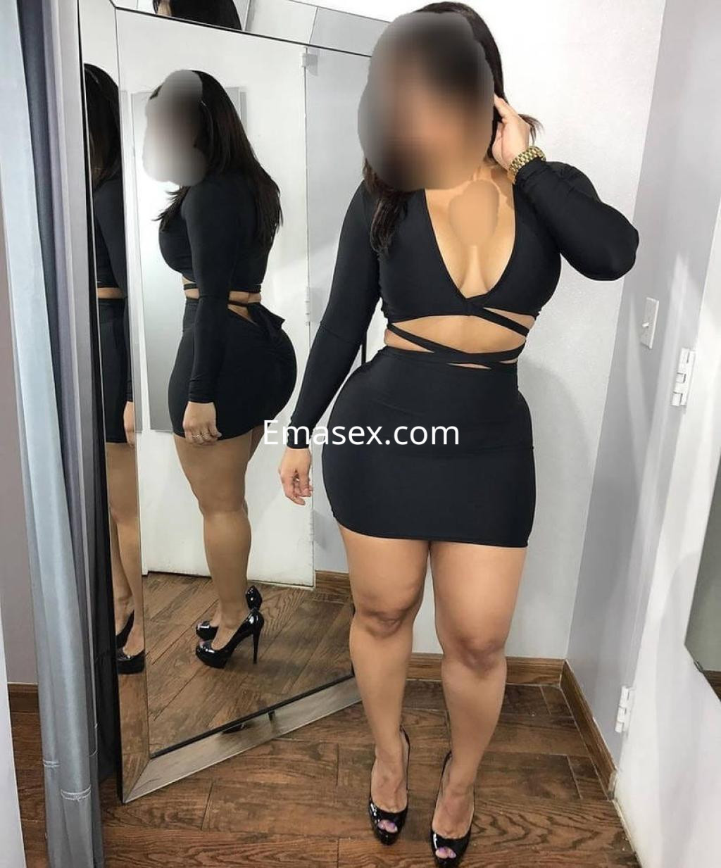 Escort en Gijón luciendo un vestido negro ajustado con cortes estratégicos y tacones altos, frente a un espejo.