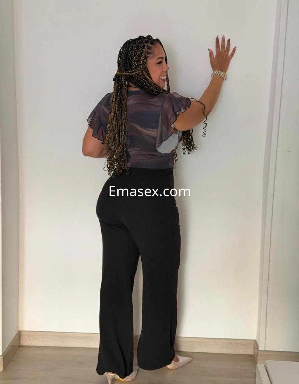 Escort en Badajoz con cabello trenzado, vistiendo blusa de manga corta y pantalones negros frente a una pared.