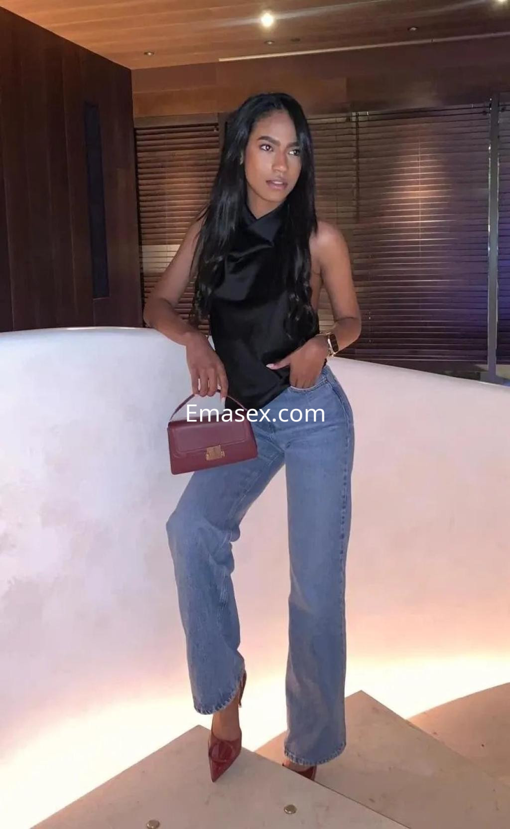 Puta de Coruña con cabello largo y lacio, viste blusa negra y jeans, posando con una bolsa roja en un ambiente elegante.