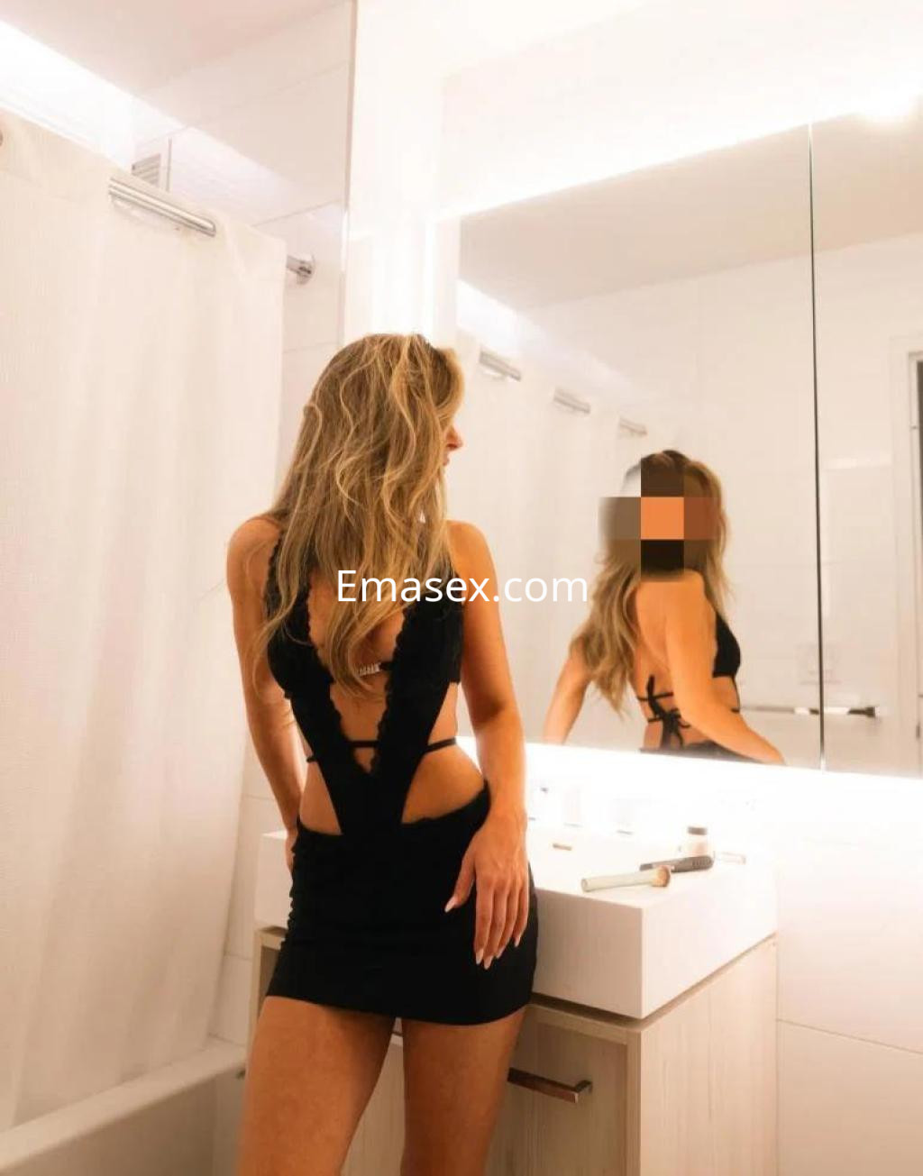 Puta posando en un baño con un vestido negro ajustado, cabello largo y rubio, reflejada en el espejo.
