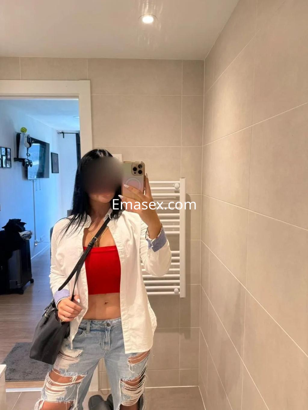 Escort en Zaragoza, posando en un baño con blusa blanca, top rojo y jeans rasgados, reflejándose en el espejo.
