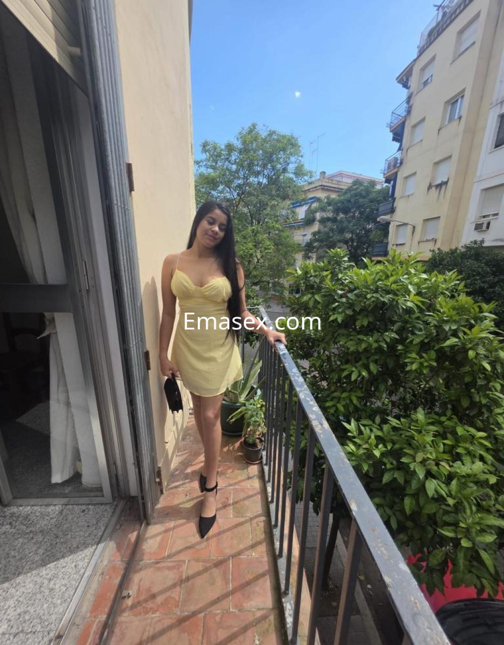 Escort en Cádiz, posando en un balcón con un vestido amarillo claro y tacones negros, rodeada de vegetación.