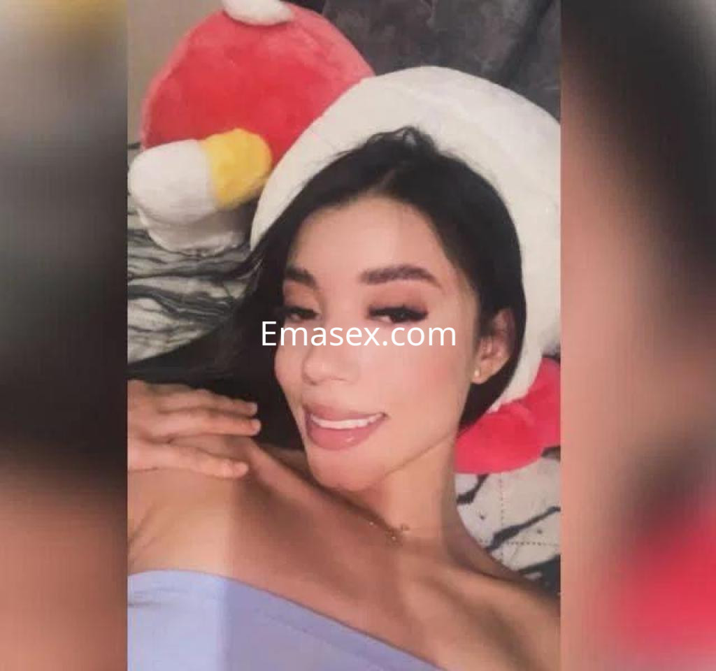 Escort en Cáceres, con cabello oscuro, posa sonriente sobre una cama rodeada de cojines coloridos.