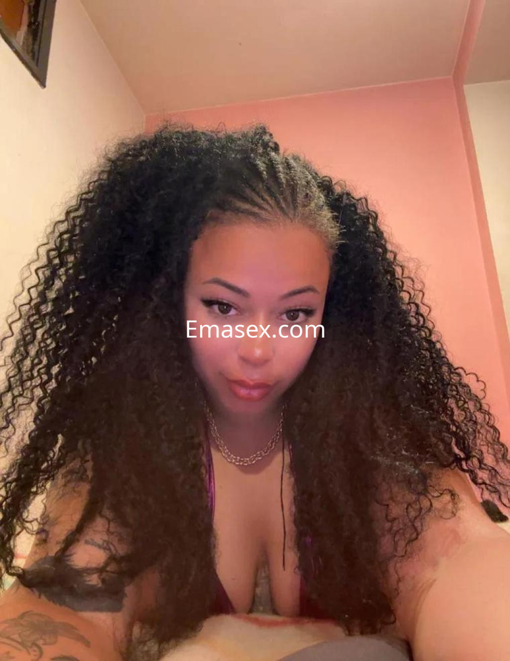 Escort con cabello rizado y voluminoso, posando en un ambiente interior con paredes rosas en Ourense.