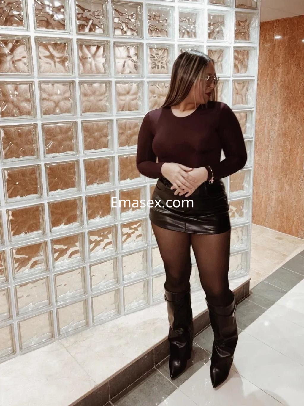 Escort en Vigo con top ajustado de color burdeos, falda corta de cuero y botas altas, posando en un ambiente moderno.