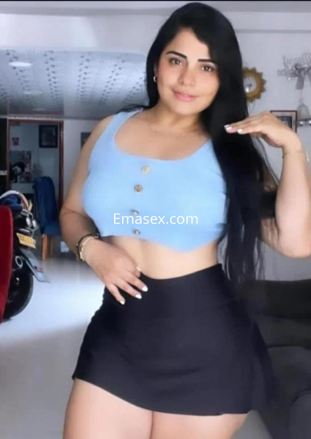 Escort en Barcelona con blusa azul y falda negra, posando con una mano en la cadera en un ambiente interior.