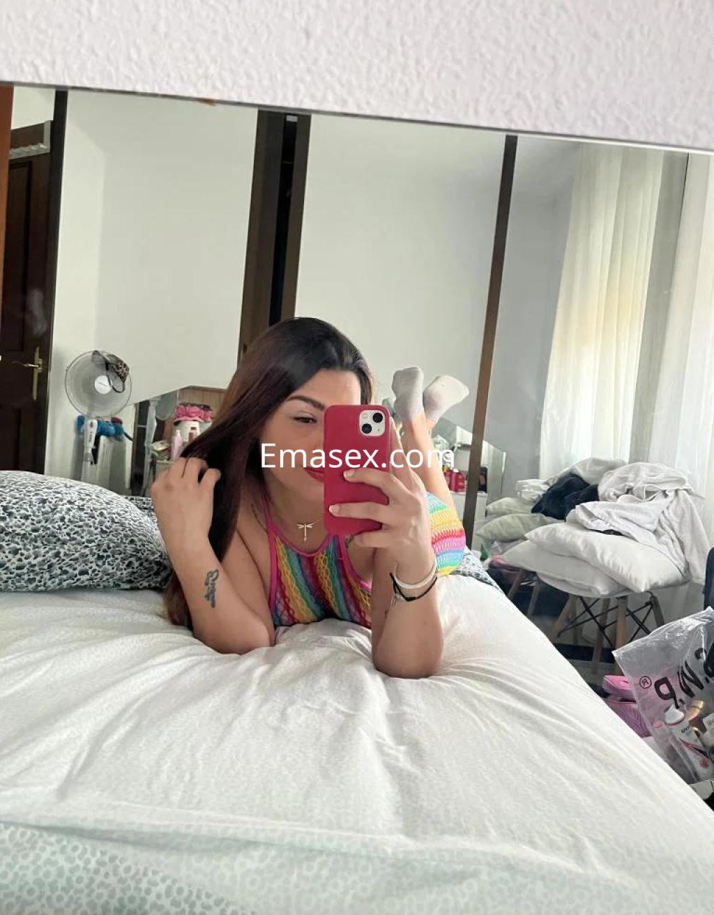 Puta con cabello largo y castaño, en una cama deshecha, posa en un ambiente sencillo con un top colorido.