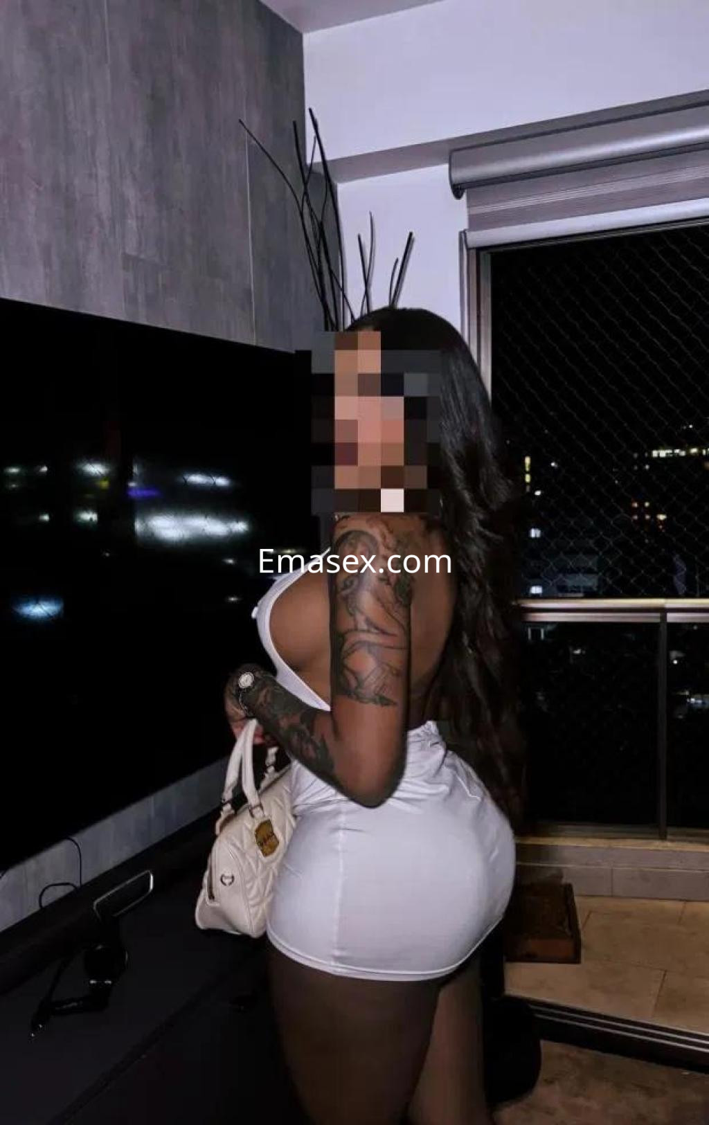 Escort en Coruña con vestido blanco ajustado, cabello oscuro y tatuajes, posando junto a una ventana.