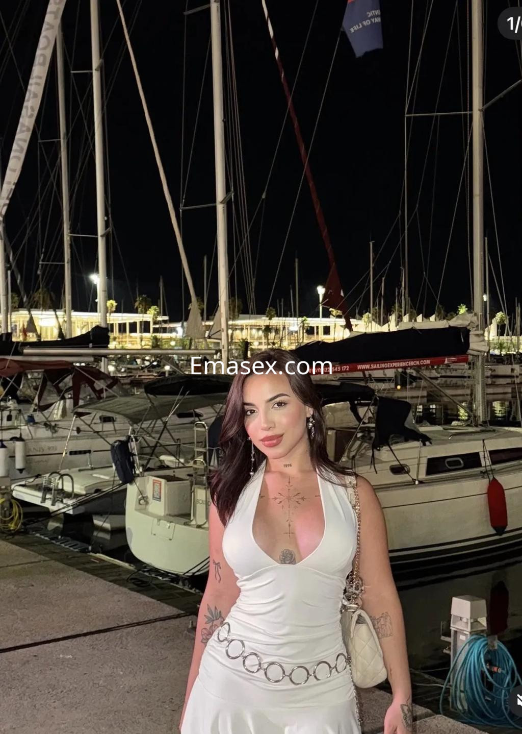 Escort en Coruña con vestido blanco, cabello oscuro y un entorno de puerto iluminado por la noche.