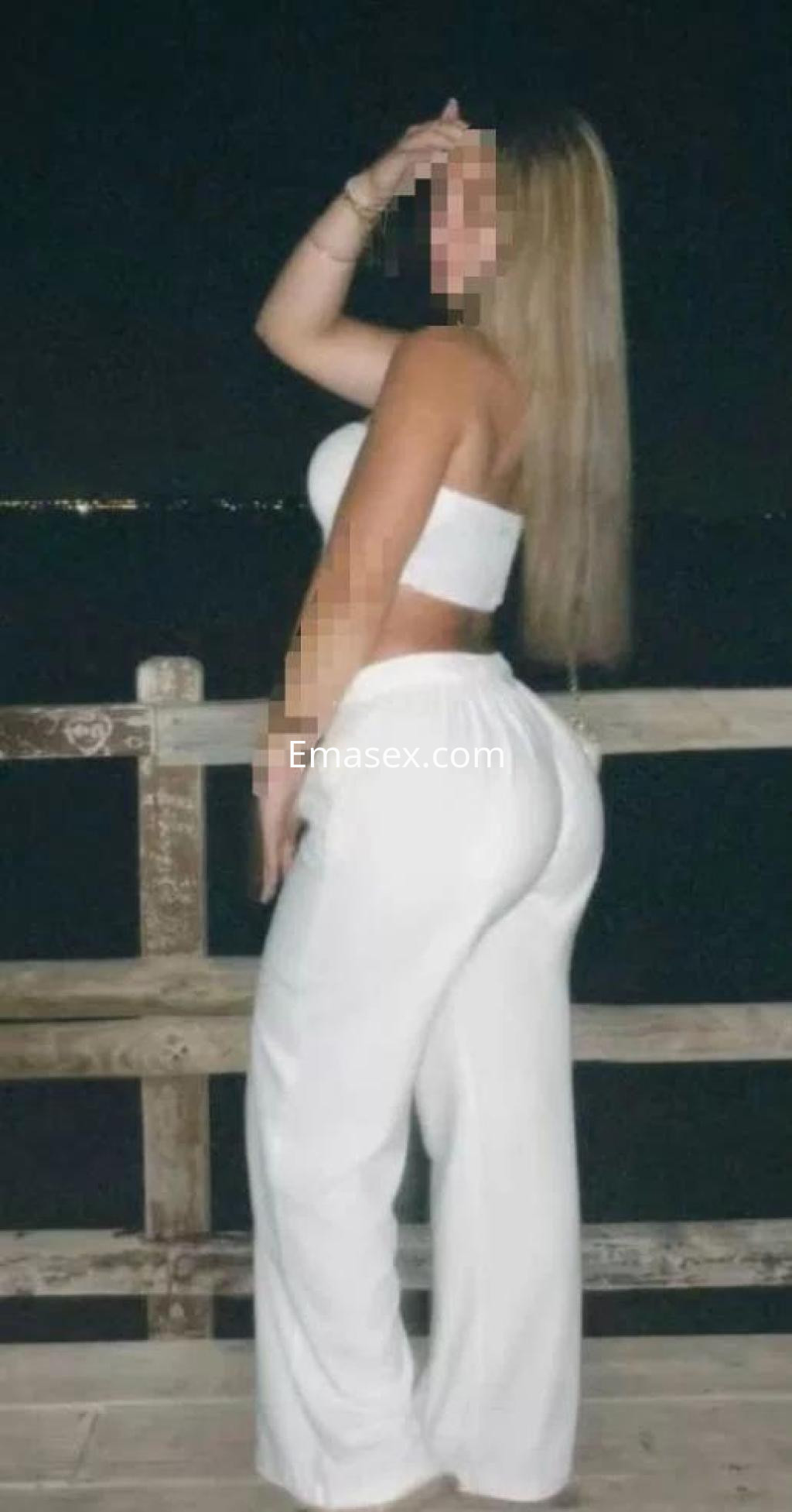 Puta posando de perfil en Toledo, vistiendo un top blanco y pantalones anchos, con cabello largo y lacio.