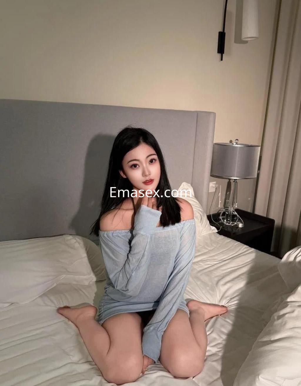 Escort en Zaragoza sentada en la cama, vistiendo un suéter gris claro que deja un hombro al descubierto.
