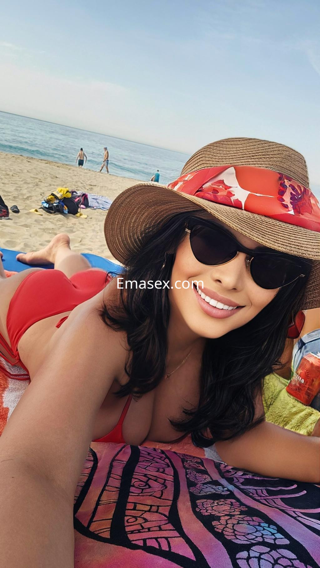 La puta en Donosti se relaja en la playa con un bikini rojo y un sombrero grande, sonriendo a la cámara.