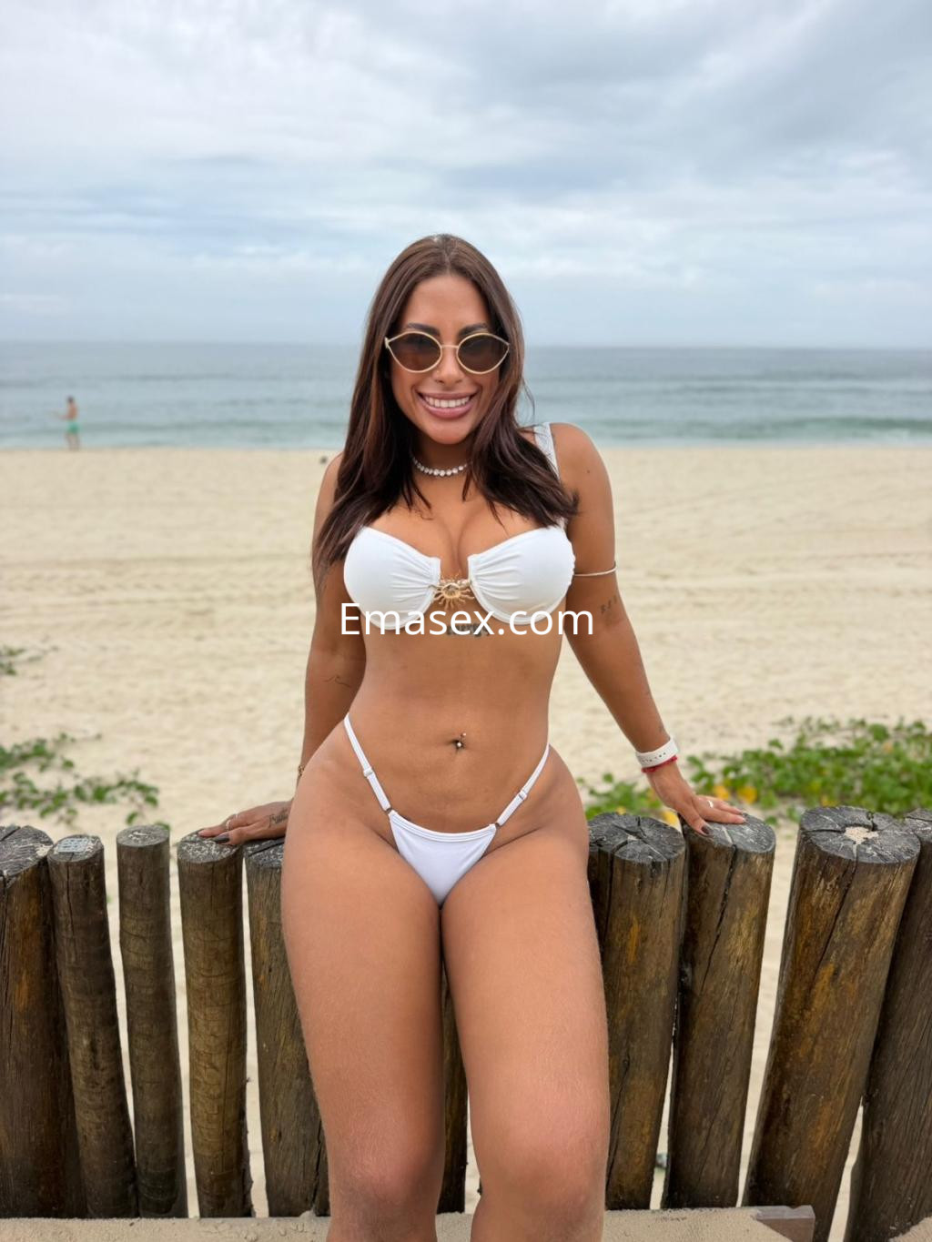 Escort en Vitoria posando en la playa con un bikini blanco y gafas de sol, sonriendo con el mar al fondo.