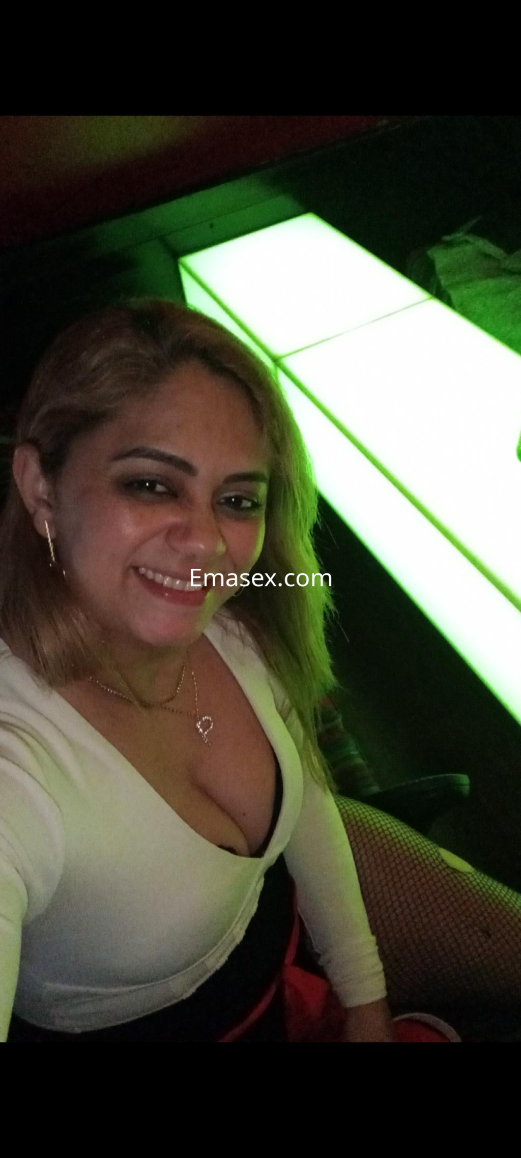 Escort en Vigo sonríe vestida con blusa blanca y falda roja, sentada en un ambiente iluminado con luces verdes.