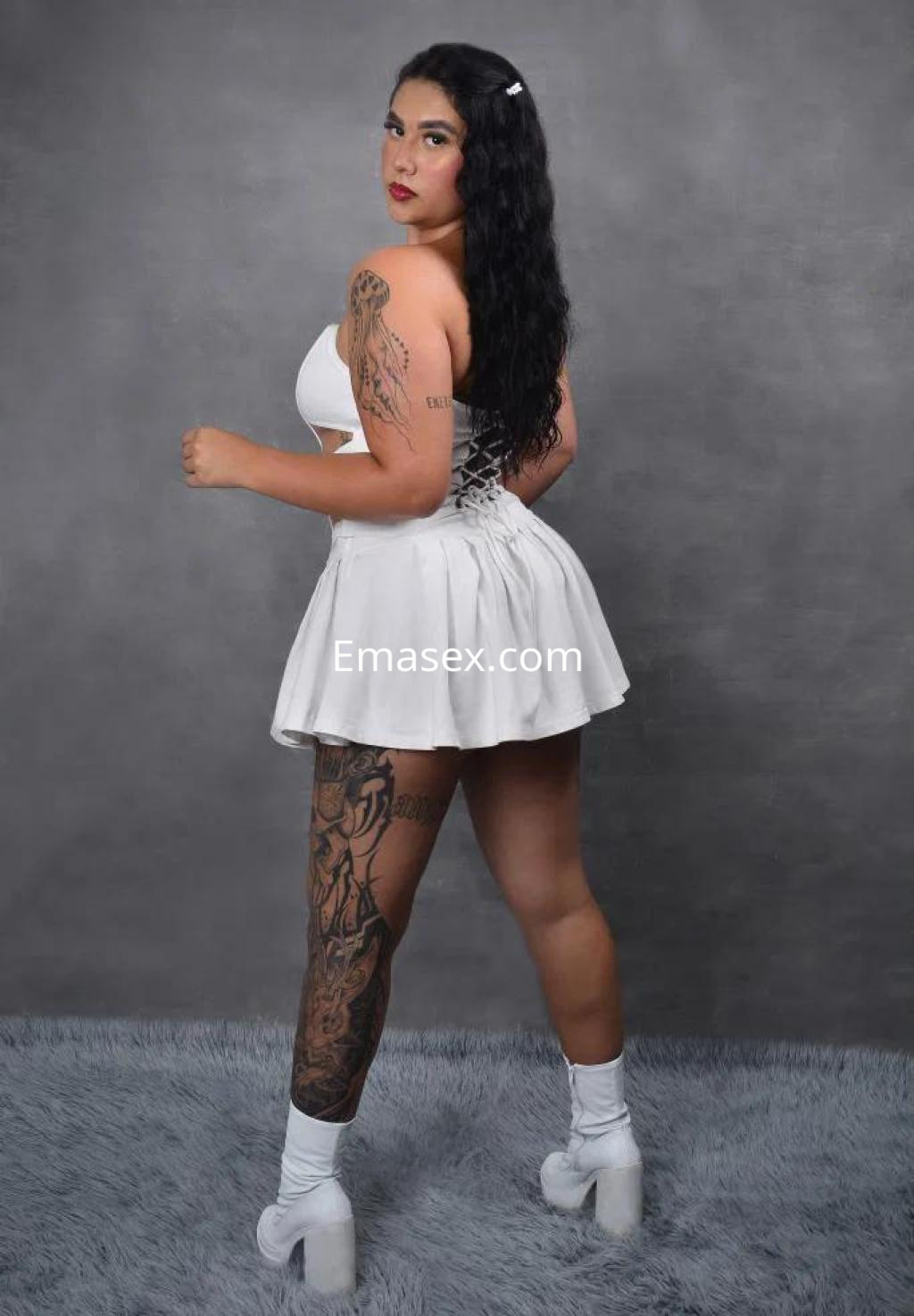 Escort en Alicante luciendo un vestido blanco corto, con tatuajes visibles y cabello largo y ondulado.