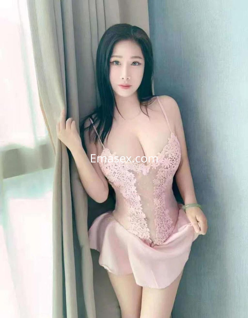 Escort en Barcelona posa junto a una cortina, luciendo un vestido rosa claro con detalles de encaje.
