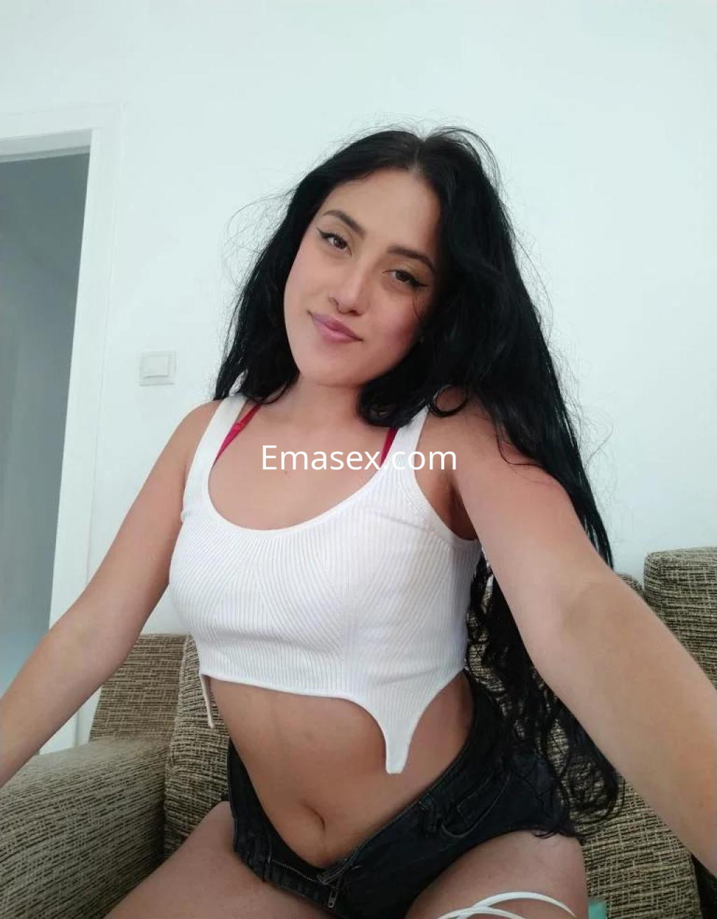 Escort en Castellón posando con una blusa blanca sin mangas y pantalones cortos, con cabello largo y oscuro.