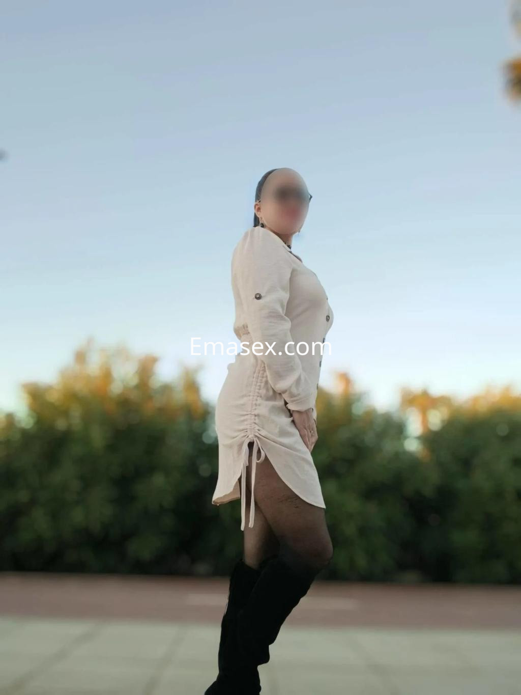 Una puta de pie en Córdoba viste un vestido claro y botas largas, posando con una mirada confiada.