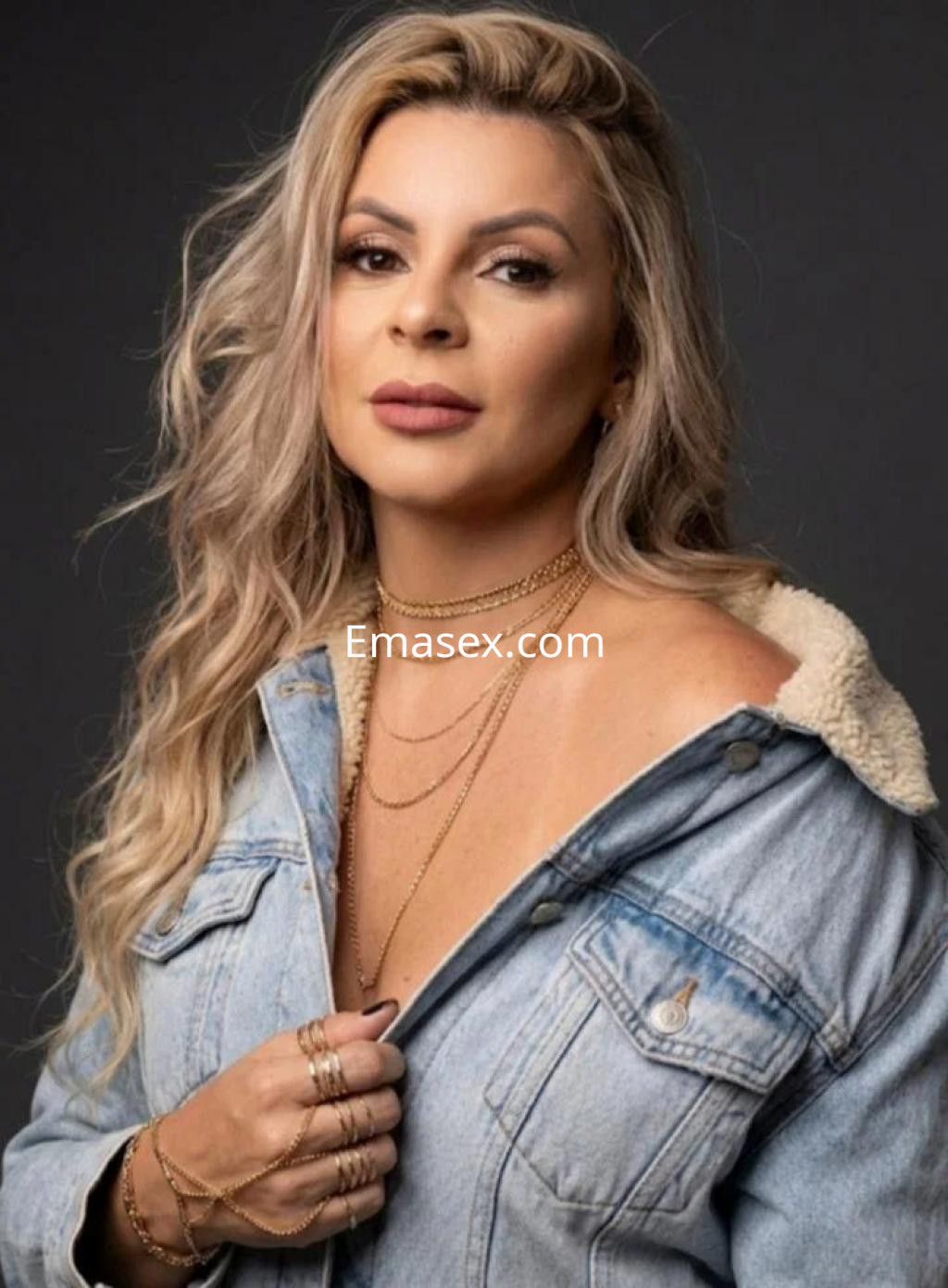 Escort en Coruña con cabello largo y rubio, viste una chaqueta de mezclilla y joyería dorada.