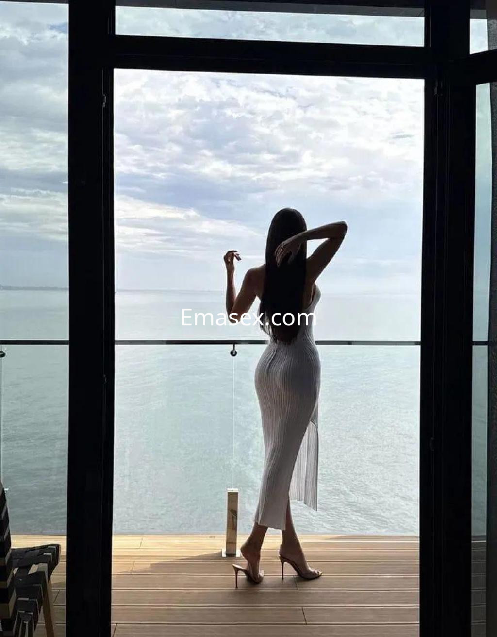 Escort en Coruña de pie junto a una ventana, vistiendo un vestido blanco ajustado que destaca su silueta.