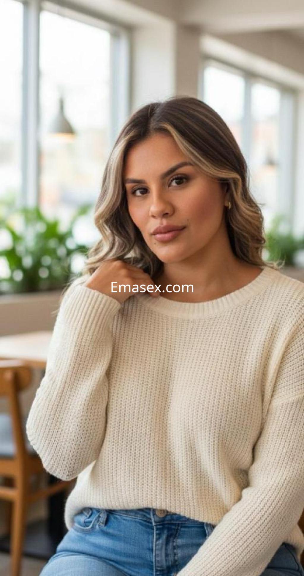 Escort en Málaga con cabello largo y ondulado, viste un suéter blanco y está sentada en un entorno luminoso.