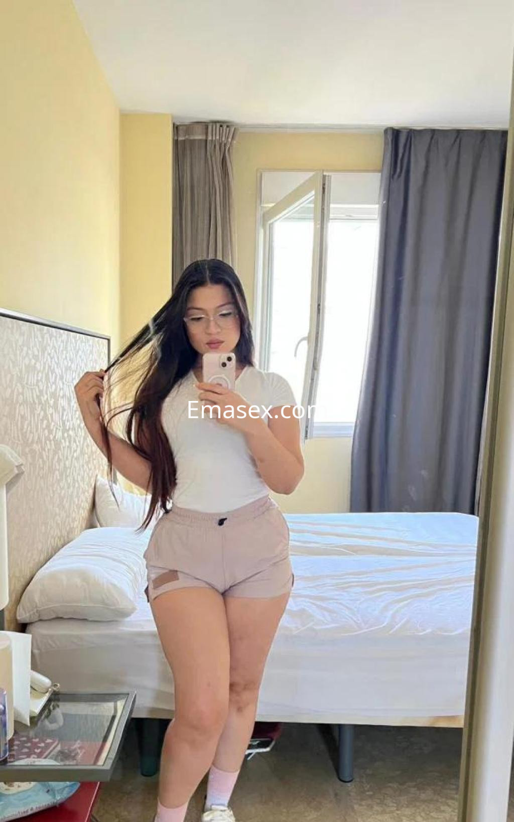 La puta en Sevilla luce una camiseta blanca y pantalones cortos, posando frente a un espejo en un dormitorio.