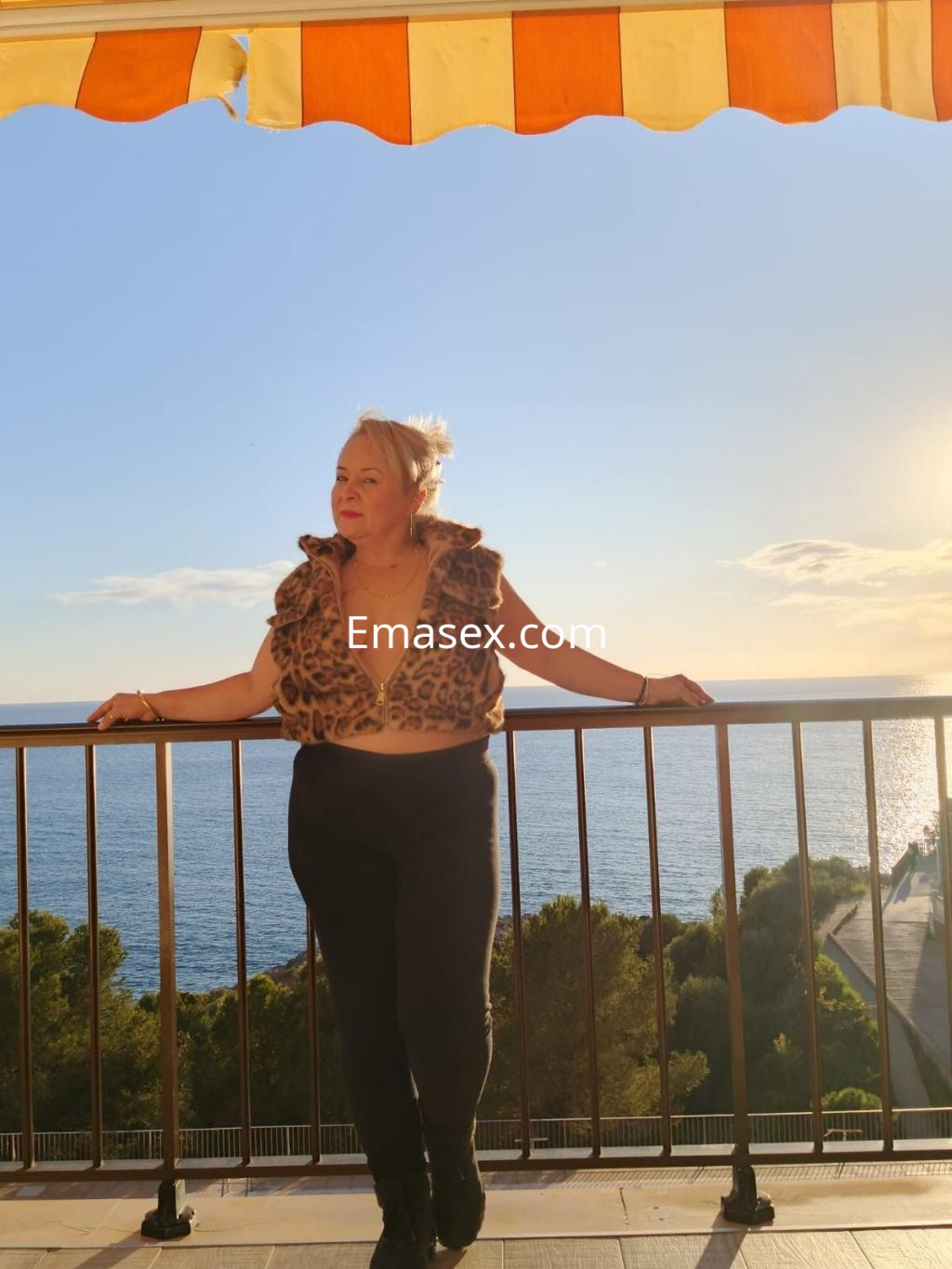 Escort en Zaragoza, con blusa de leopardo y pantalones negros, posando en un balcón con vista al mar.