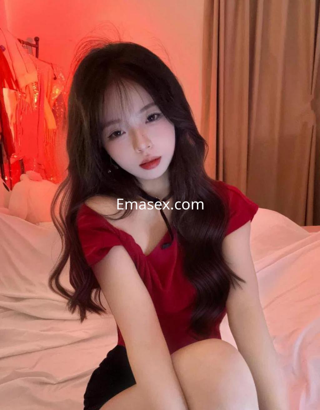 Escort en Zaragoza con cabello largo y ondulado, vistiendo una blusa roja y sentada en una cama con fondo suave.