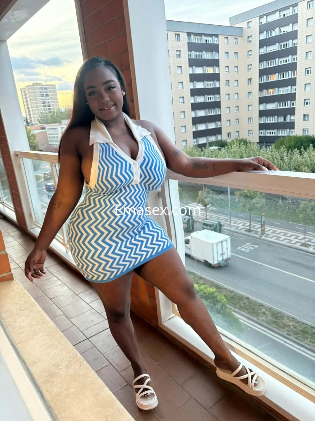 Escort en Zaragoza posando junto a una ventana, vistiendo un vestido de rayas azul y blanco y sandalias blancas.