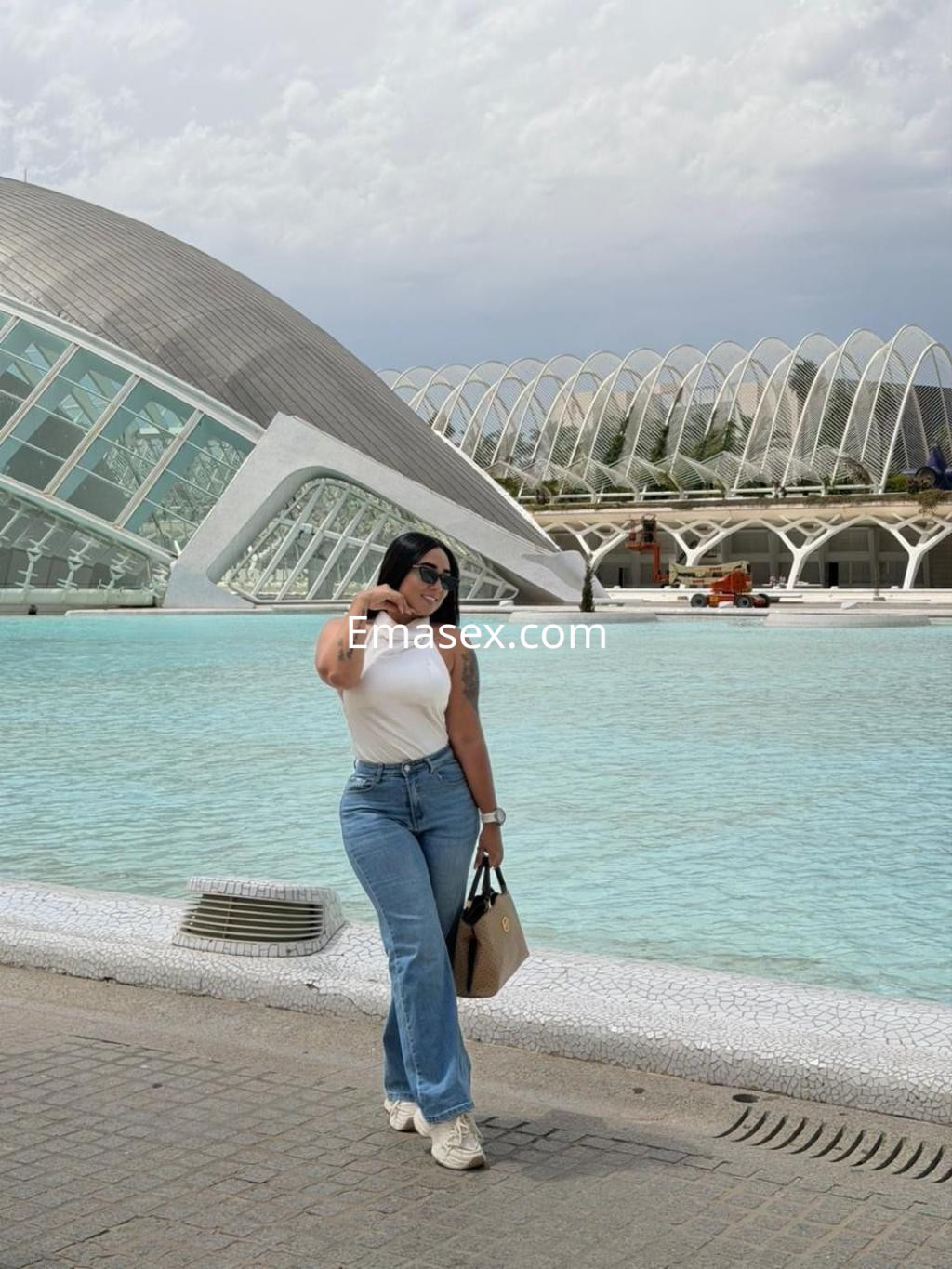 Puta en Alicante posa frente a un moderno edificio, vistiendo una camiseta blanca y jeans, con cabello oscuro y lacio.