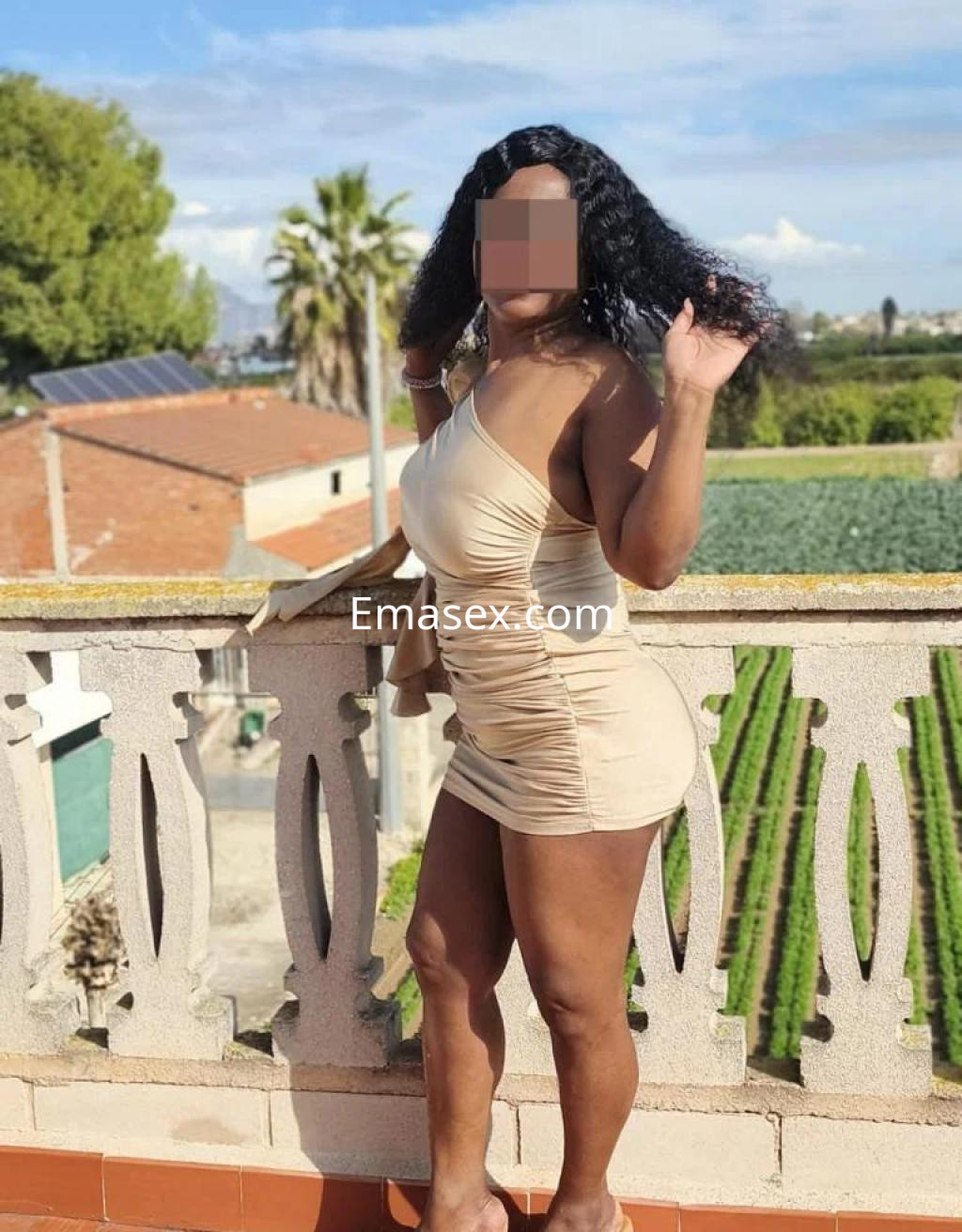 Escort en Alicante con vestido ajustado beige, cabello rizado y fondo de paisaje con palmeras.