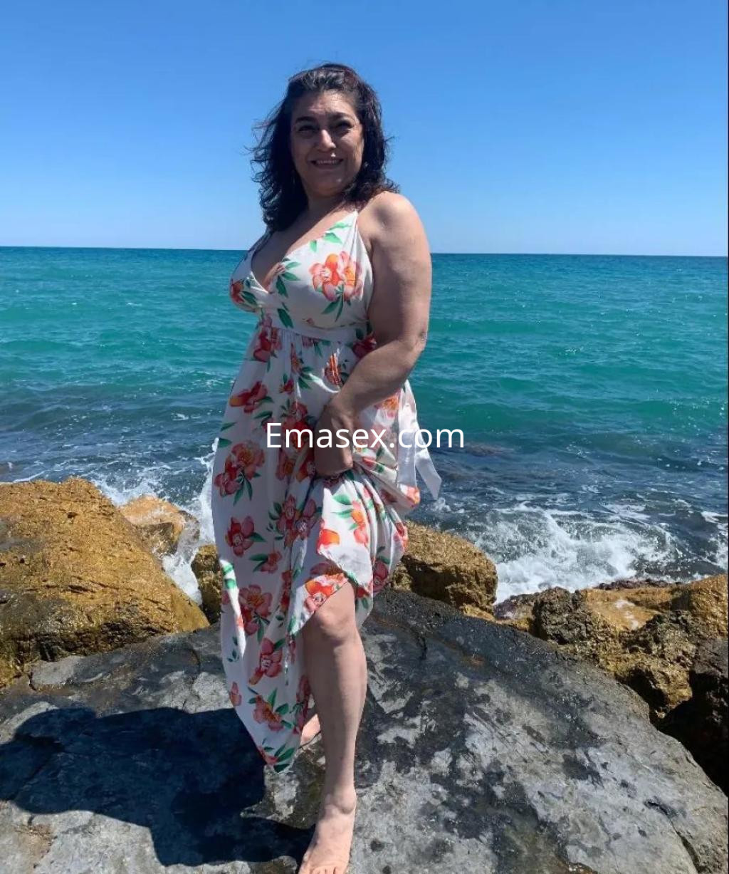 Escort en Alicante luciendo un vestido floral, de pie sobre rocas junto al mar, con el cabello suelto y ondulado.