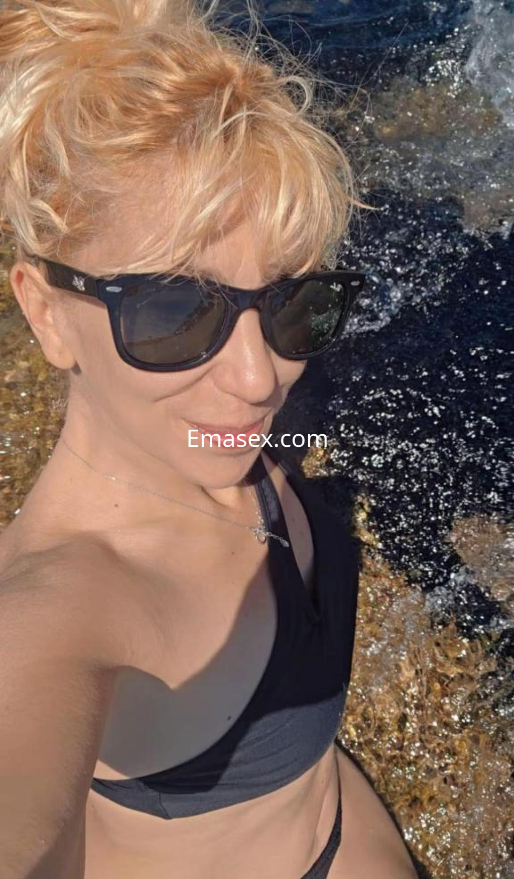 Escort en Barcelona posando en la playa con gafas de sol, cabello rubio rizado y bikini negro.