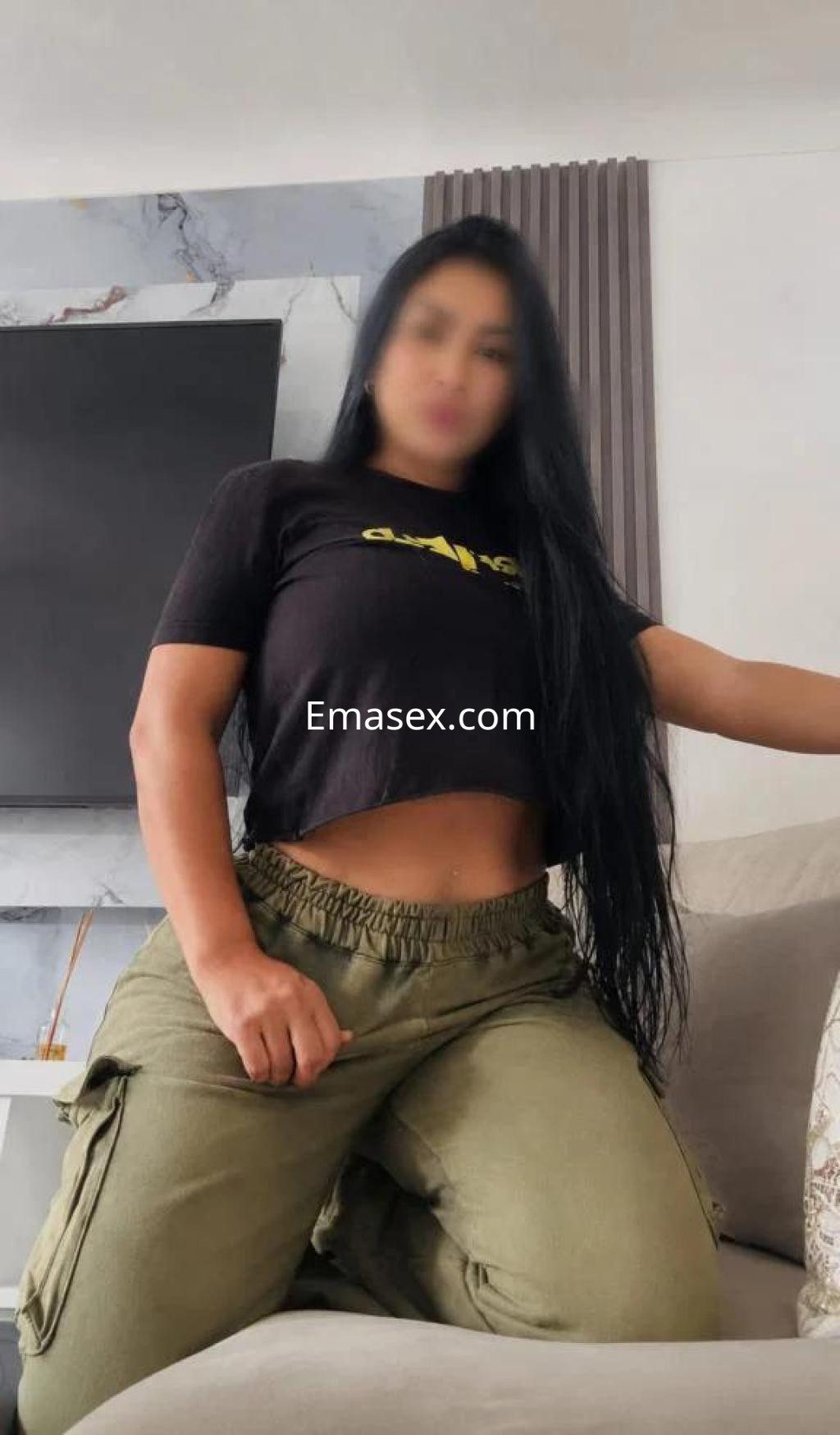 La puta viste una camiseta negra ajustada y pantalones cargo verdes, posando en un salón moderno en Barcelona.