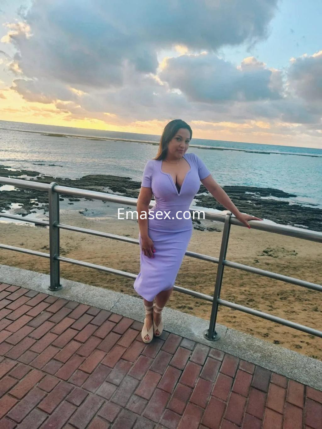 Escort posando en un vestido lila ajustado junto al mar en Cádiz, con un fondo de atardecer.