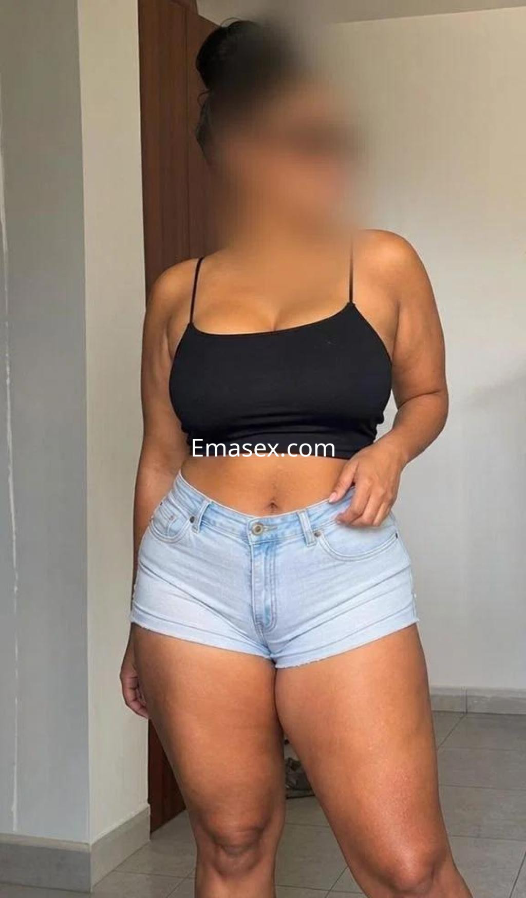 Puta de Castellón viste una blusa negra sin mangas y shorts de mezclilla, mostrando su figura en un entorno interior.