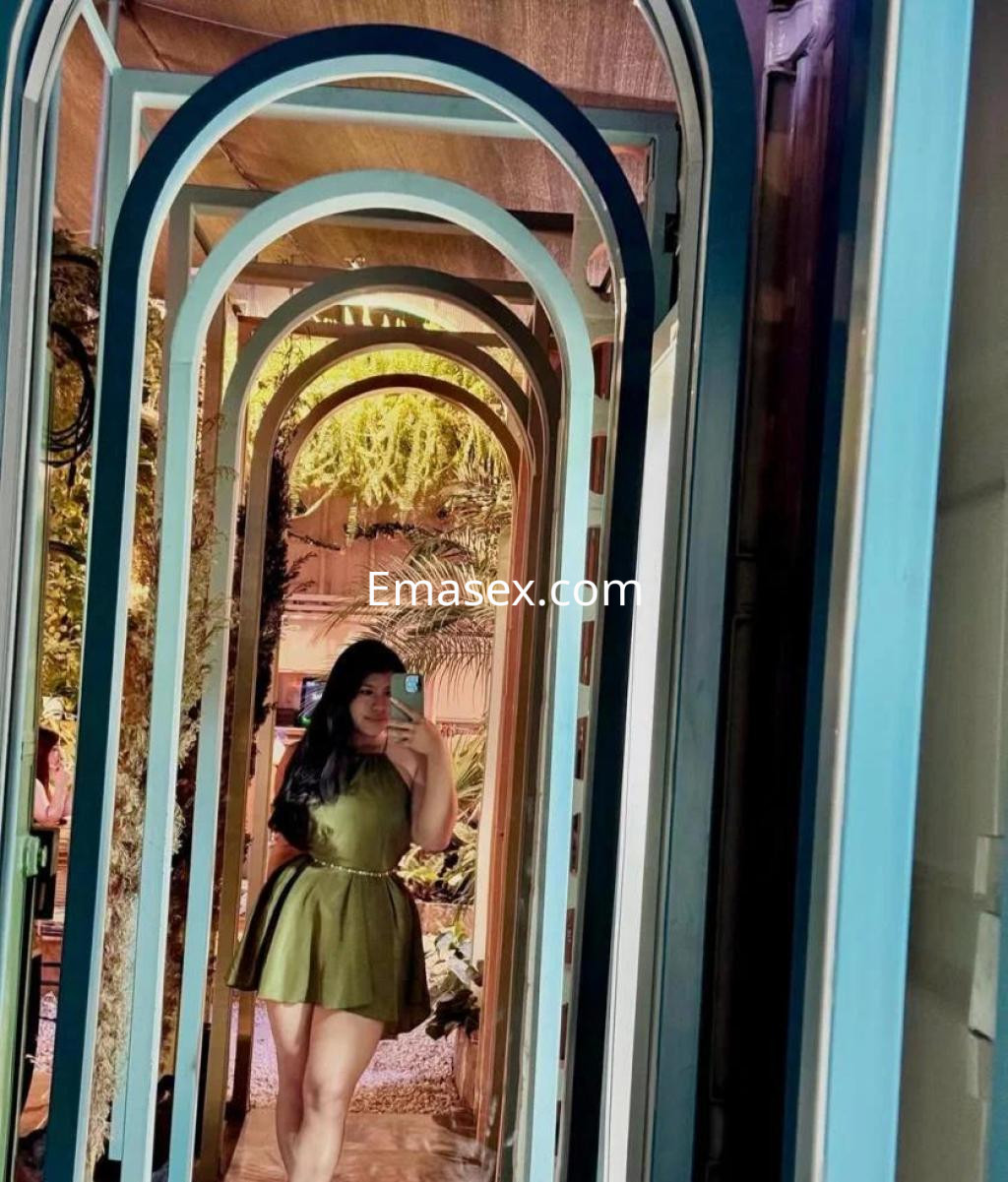 Escort en Granada posando en un pasillo con arcos decorativos, vistiendo un vestido verde y sosteniendo un teléfono.
